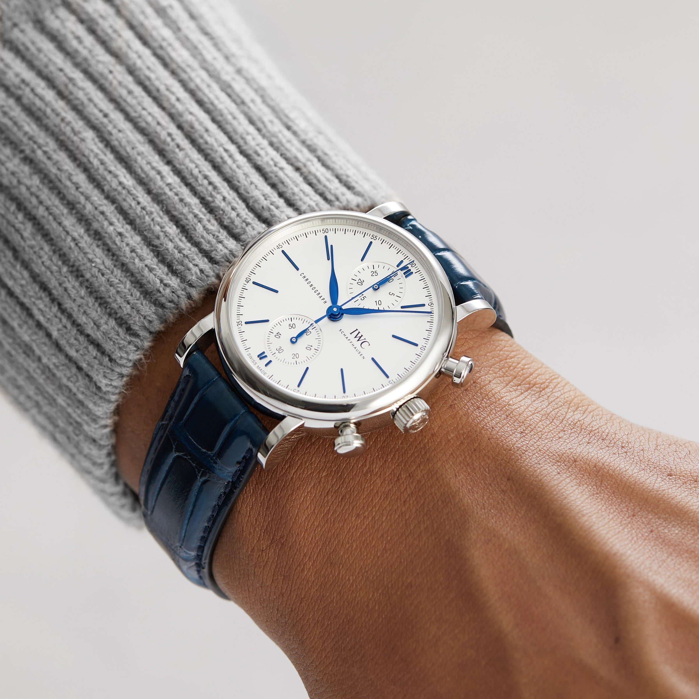 A person wearing the IWC Schaffhausen Portofino Chronograph, featuring a silver stainless steel case, white dial with blue markers, and a blue alligator leather strap.