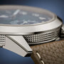Close-up of the Patek Philippe Complications 5328G-001 watch case, featuring textured metal sides and a brown leather strap with white stitching.