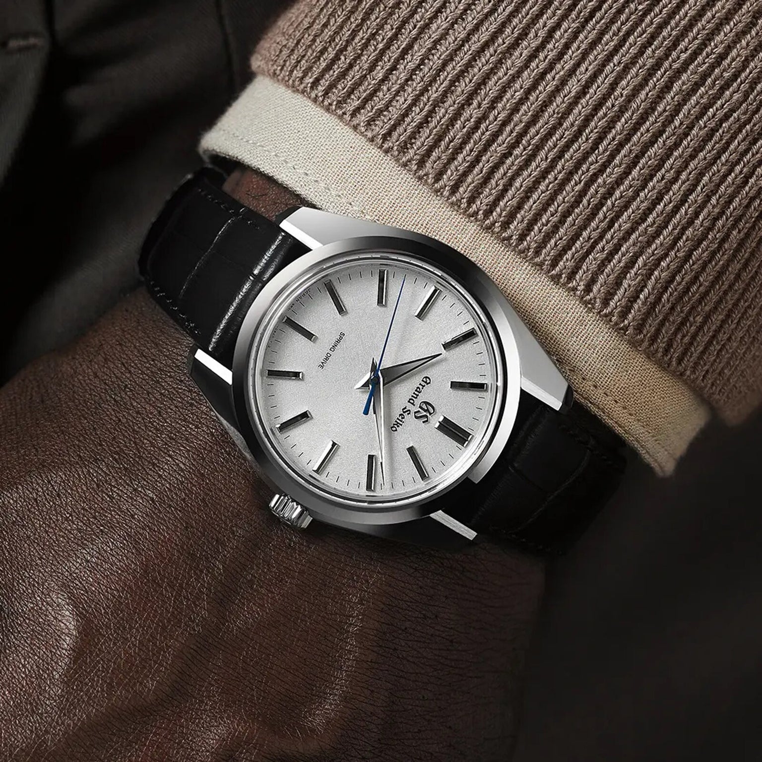 A close-up of a wrist wearing the Grand Seiko Spring Drive Heritage watch with a silver case, black leather strap, white textured dial, and blue second hand, paired with a brown leather glove and beige sweater.