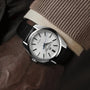 A close-up of a wrist wearing the Grand Seiko Spring Drive Heritage watch with a silver case, black leather strap, white textured dial, and blue second hand, paired with a brown leather glove and beige sweater.