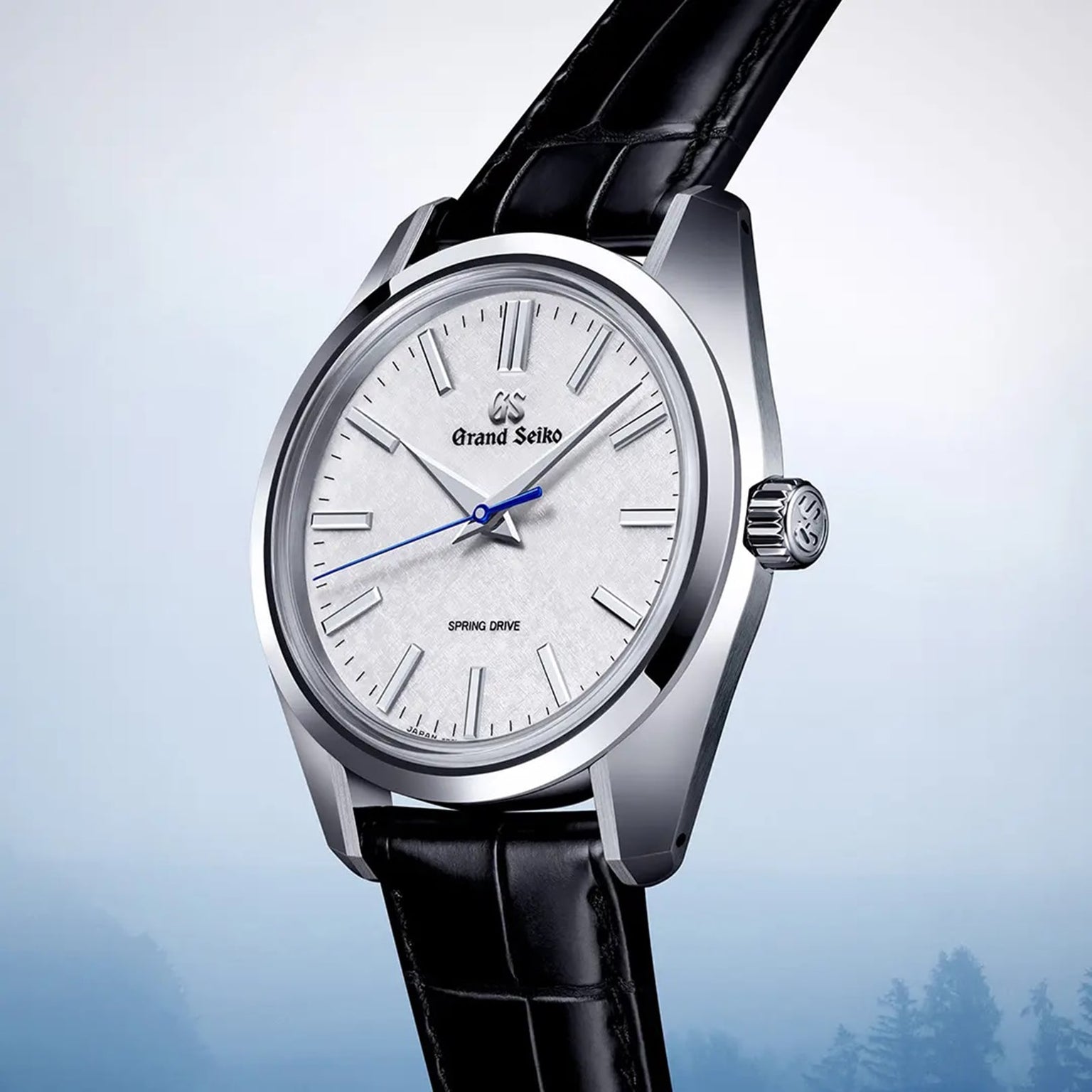 The Grand Seiko Spring Drive Heritage wristwatch, featuring a silver dial, black leather strap, and blue second hand, is displayed against a light blue gradient background to highlight its precise Spring Drive movement.