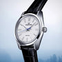 The Grand Seiko Spring Drive Heritage wristwatch, featuring a silver dial, black leather strap, and blue second hand, is displayed against a light blue gradient background to highlight its precise Spring Drive movement.