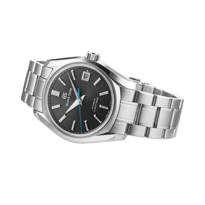 The Grand Seiko Hi-Beat Heritage, a stainless steel watch inspired by Jodogahama beach, features a black textured dial with silver hour markers, blue second hand, date at 3 o’clock, and metal link bracelet—pictured against a white background.