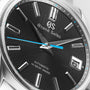 Close-up of the Grand Seiko Hi-Beat Heritage wristwatch, featuring a textured black dial, silver hour markers, blue second hand, and date display at 3 o’clock, inspired by the serene beauty of Jodogahama beach.