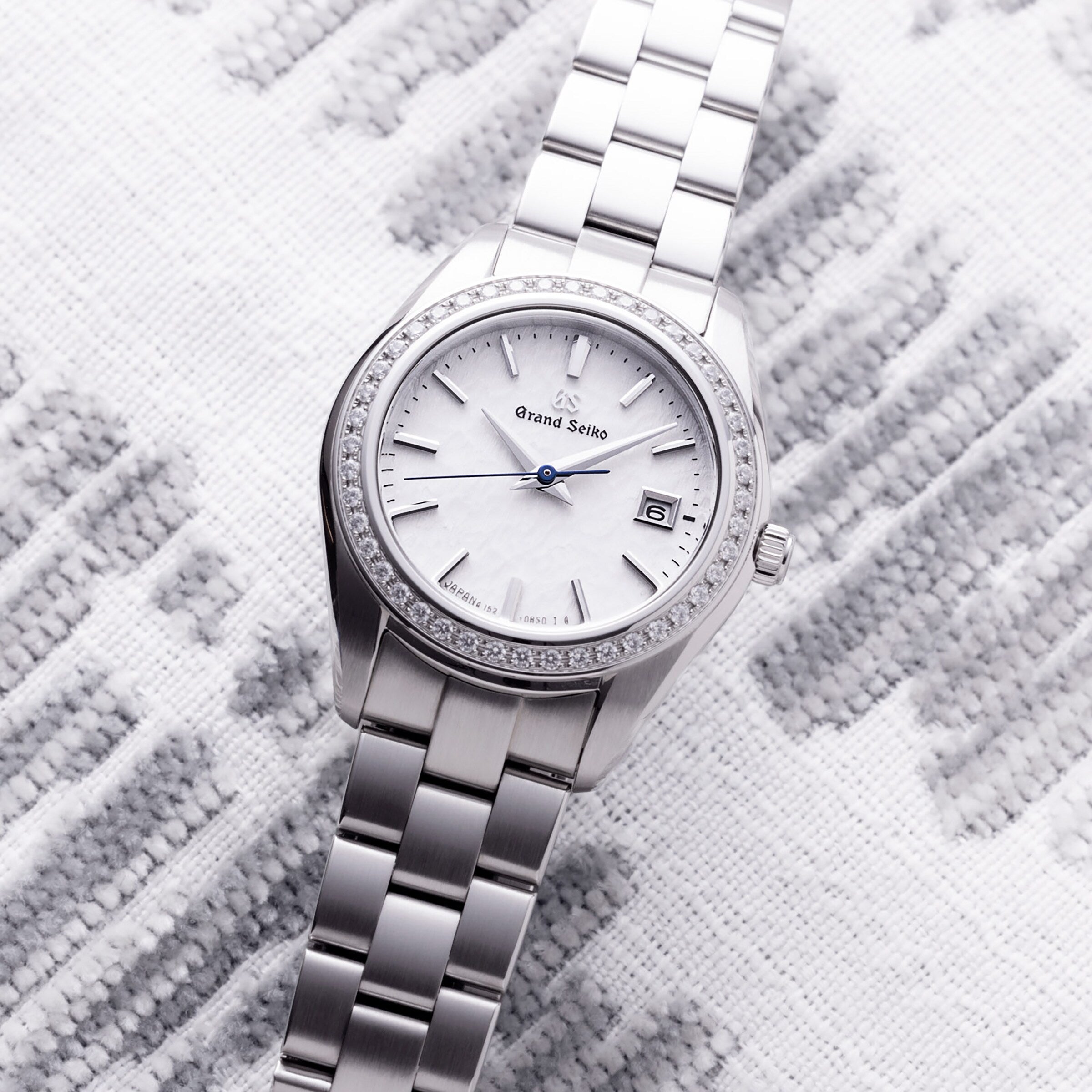 The Grand Seiko Ladies Heritage wristwatch features a silver case, snowflake dial, date display, blued steel seconds hand, and 44 diamonds on the bezel, displayed on a textured white and gray surface.