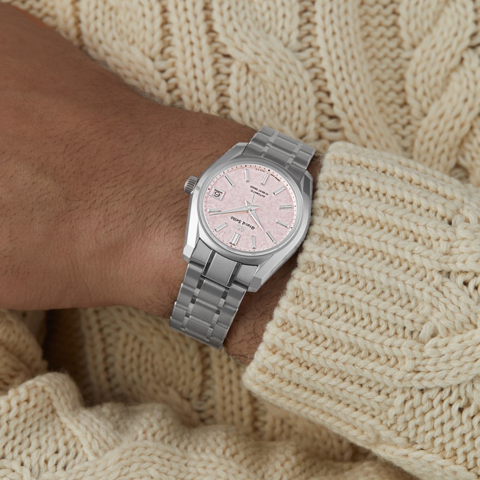 A person wears the Grand Seiko Hi-Beat Heritage watch, featuring a silver case, light pink dial, and metal link band, styled with a cream knit sweater for a refined Heritage Collection-inspired look.