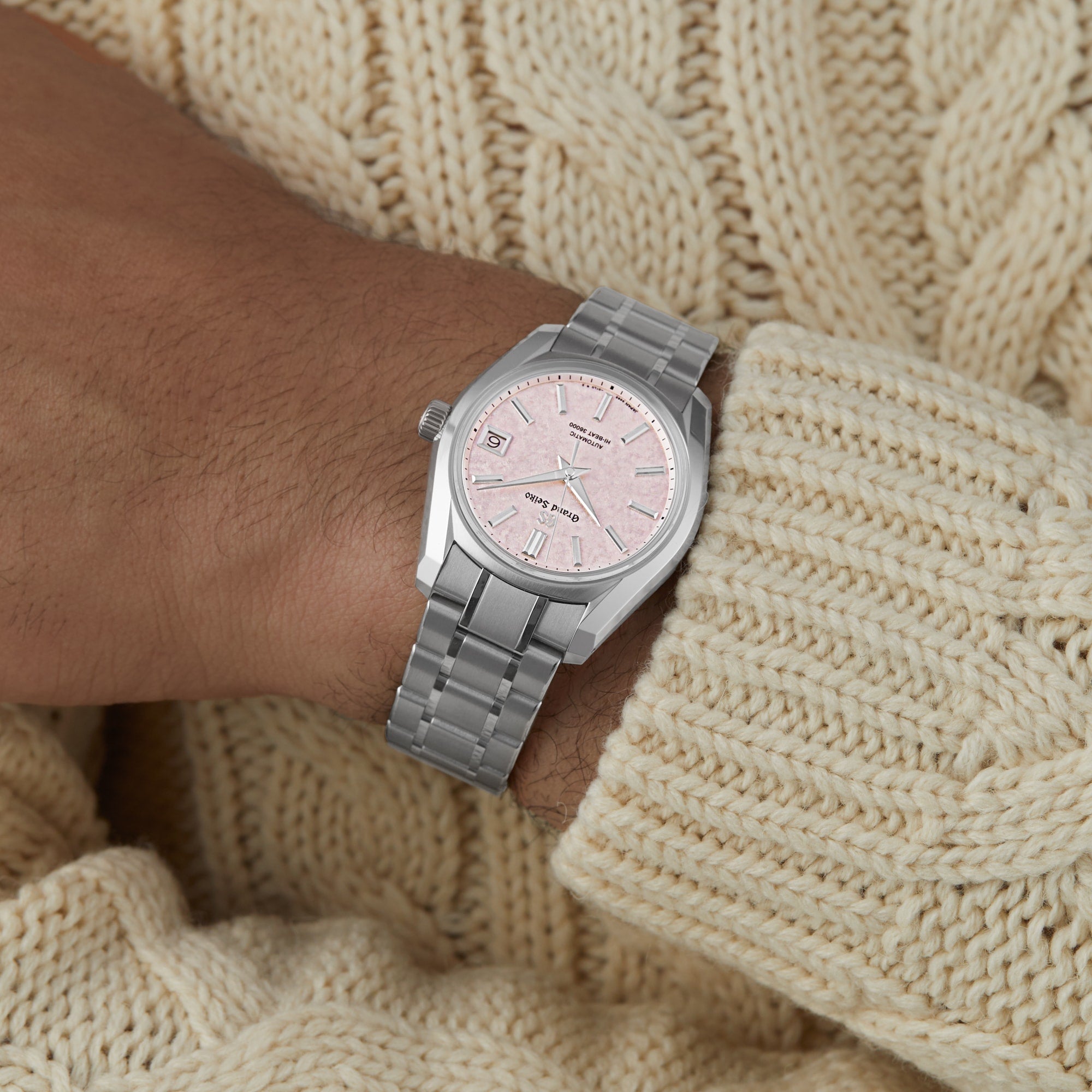 A person wears the Grand Seiko Hi-Beat Heritage watch, featuring a silver case, light pink dial, and metal link band, styled with a cream knit sweater for a refined Heritage Collection-inspired look.