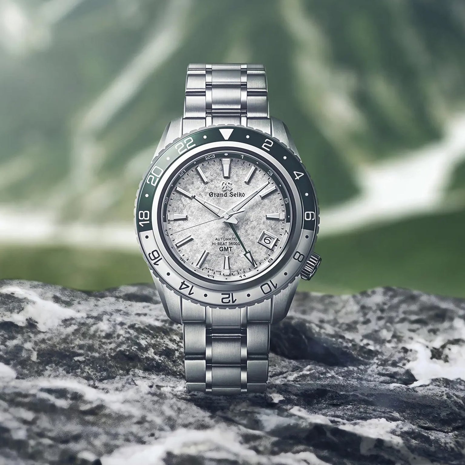 The Grand Seiko Hi-Beat GMT Sport wristwatch, featuring a textured silver dial and green bezel, is displayed on a rock with blurred mountains in the background.