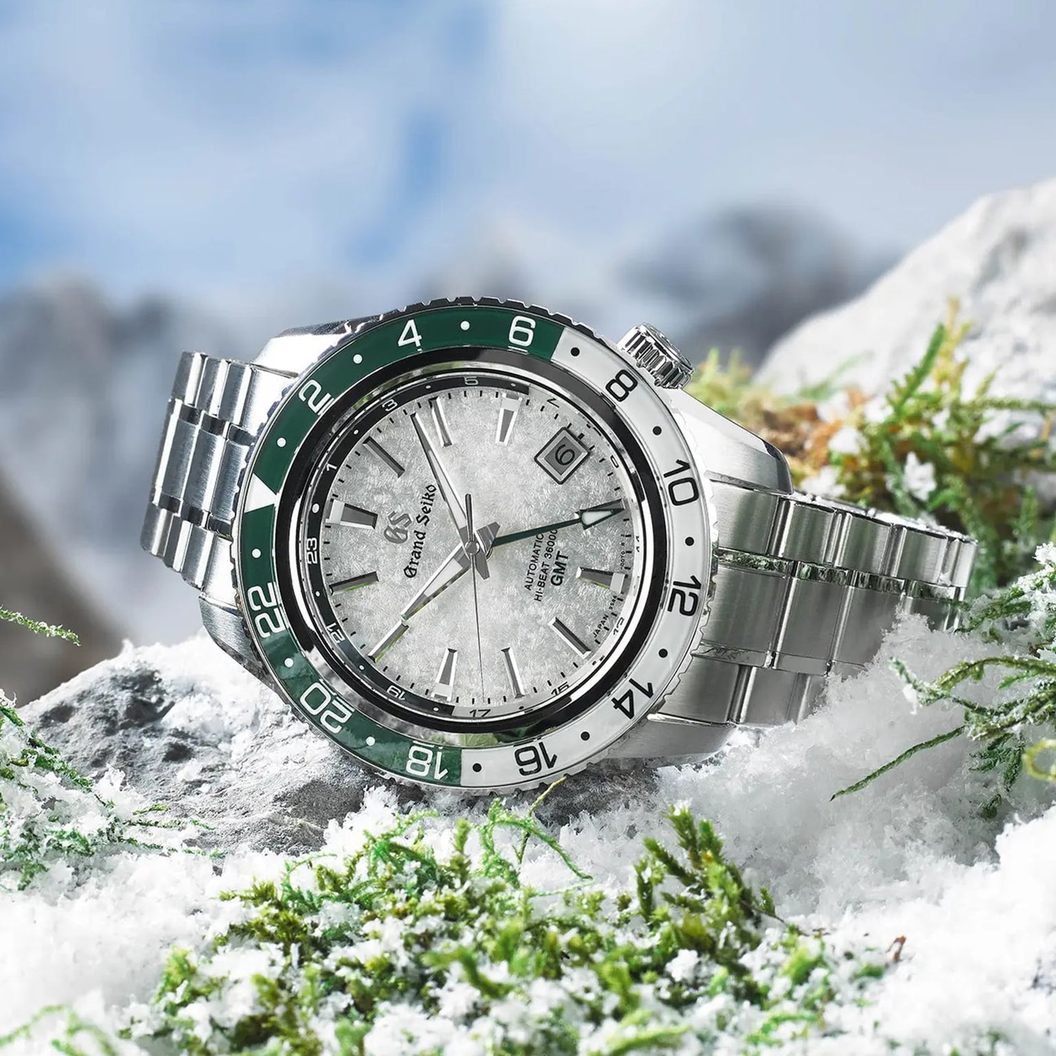 The Grand Seiko Hi-Beat GMT Sport, a stainless steel watch with a textured dial and green and silver bezel, is displayed outdoors on snow-dusted rocks with blurred mountains in the background.