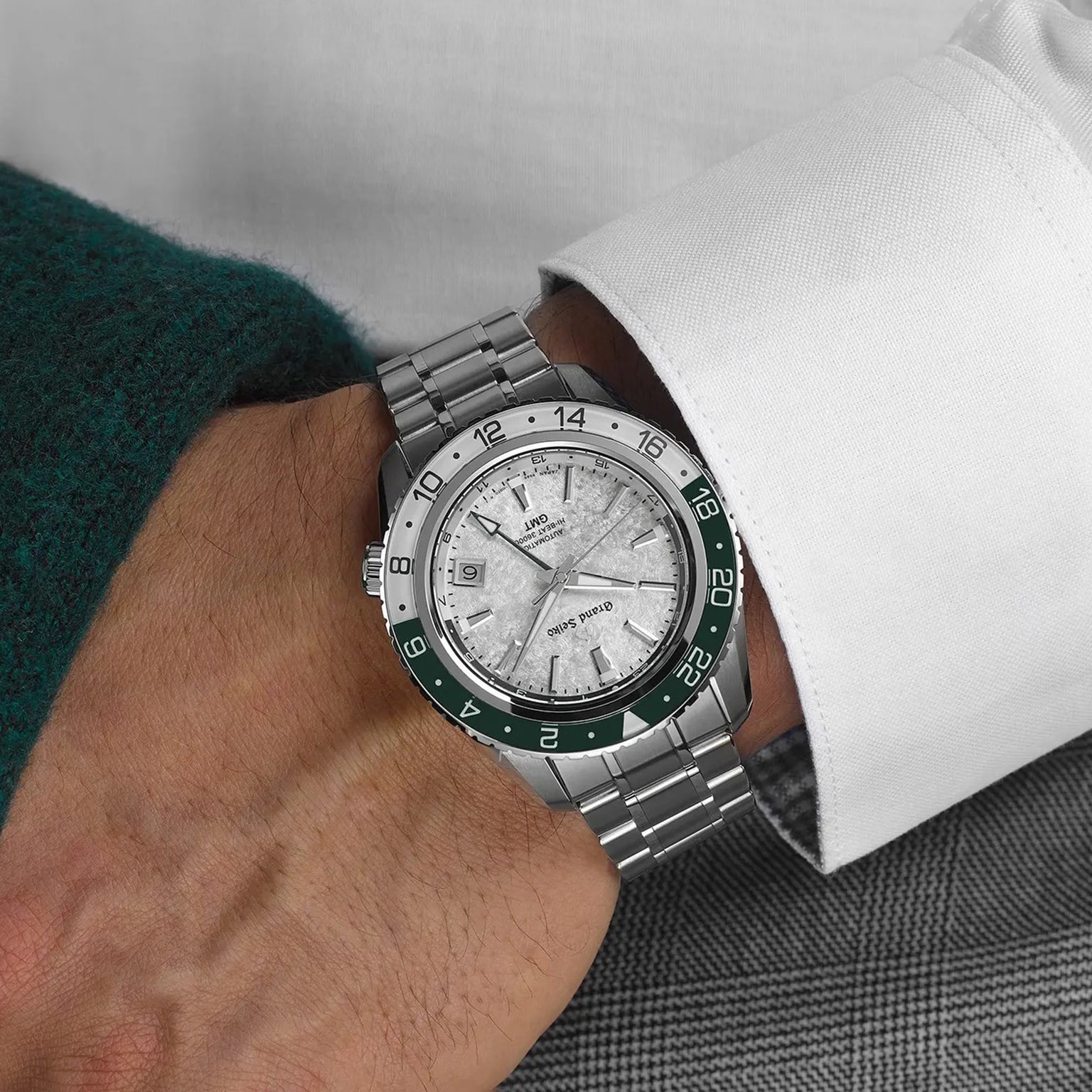 A person wearing the Grand Seiko Hi-Beat GMT Sport, featuring a green bezel and textured dial, styled with a white shirt and green sweater.