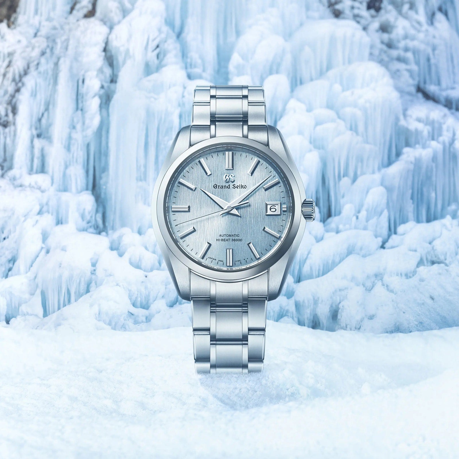 The Grand Seiko Hi-Beat Heritage wristwatch with a frosted light blue dial stands upright amid icy snow formations, highlighting the elegance and resilience of this mechanical timepiece.