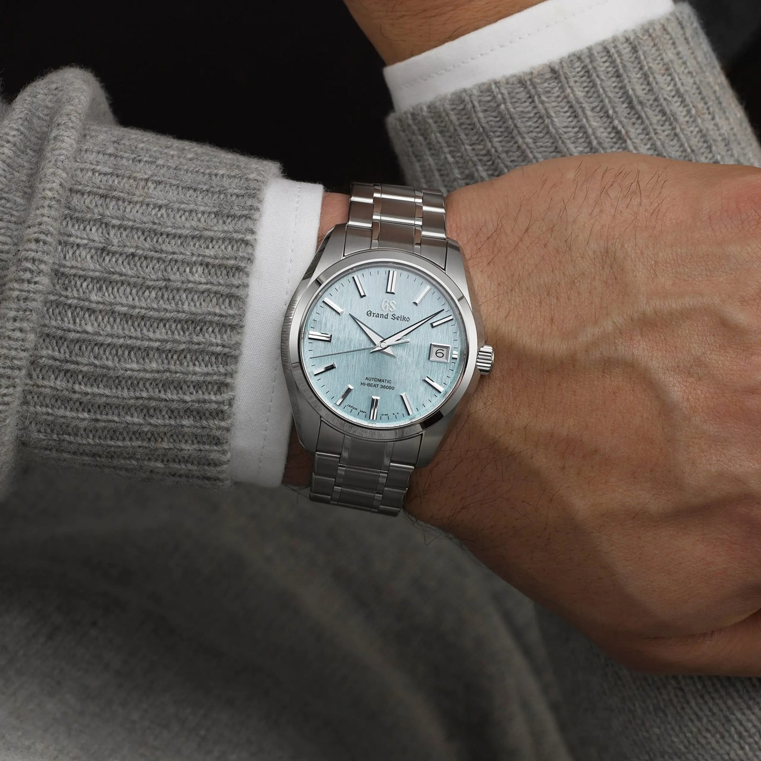 Someone wears a Grand Seiko Hi-Beat Heritage watch with a light blue textured dial, paired with a long-sleeved grey sweater and a visible white shirt cuff.
