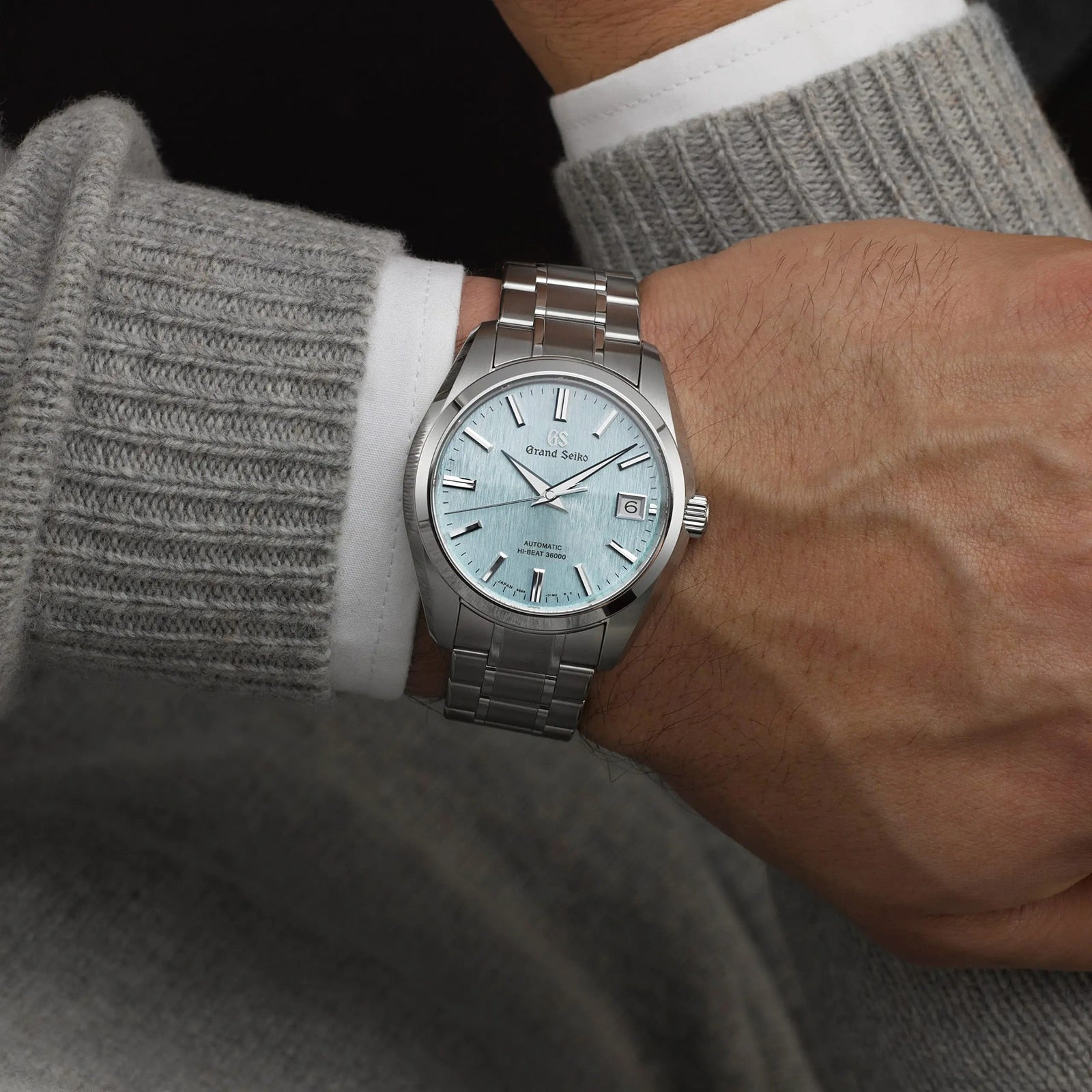Someone wears a Grand Seiko Hi-Beat Heritage watch with a light blue textured dial, paired with a long-sleeved grey sweater and a visible white shirt cuff.