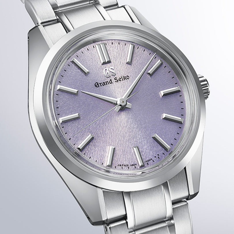 The Grand Seiko Manual SBGW323 Heritage wristwatch features a silver metal band, a textured light purple dial inspired by Studio Shizukuishi, Zaratsu-polished surfaces, plus silver hour markers and hands.