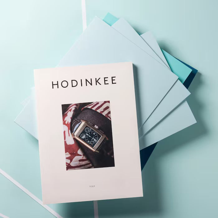 HODINKEE Magazine, Volume 9 by Hodinkee rests on light blue and teal folders, showcasing a wristwatch on the cover and celebrating horology against a light blue background.