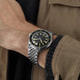 A person wearing the DOXA Sub 200 Sharkhunter divers watch, featuring a black and yellow dial with a unidirectional rotating bezel, paired with a brown jacket and black sweater.