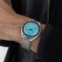 A person wearing the DOXA Sub 200 Aquamarine, a divers watch featuring a bright turquoise face and silver 316L stainless steel band, shown on their left wrist with a dark jacket and grey sweater.