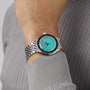 A person wearing the DOXA Sub 200 Aquamarine divers watch, featuring a turquoise face, silver hour markers, and a 316L stainless steel strap, paired with a light gray ribbed long-sleeve shirt.
