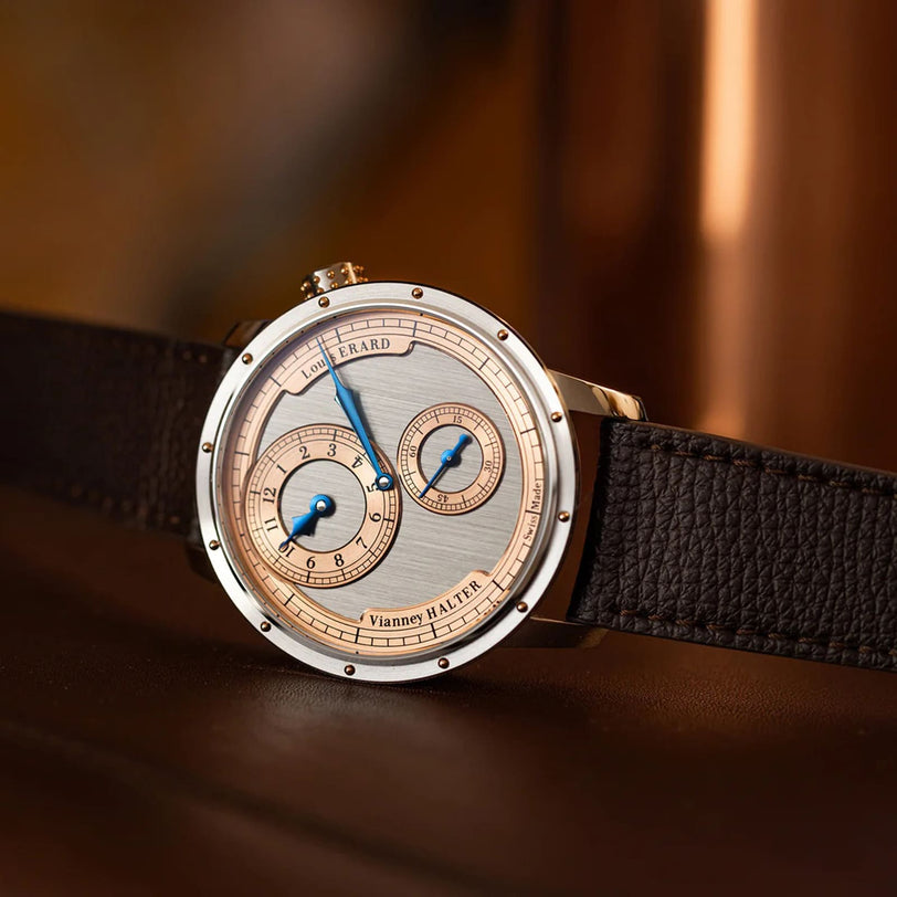The Louis Erard Le Régulateur Louis Erard x Vianney Halter II is a luxury wristwatch featuring a brown leather strap and dual subdials, shown against a warm-toned background.
