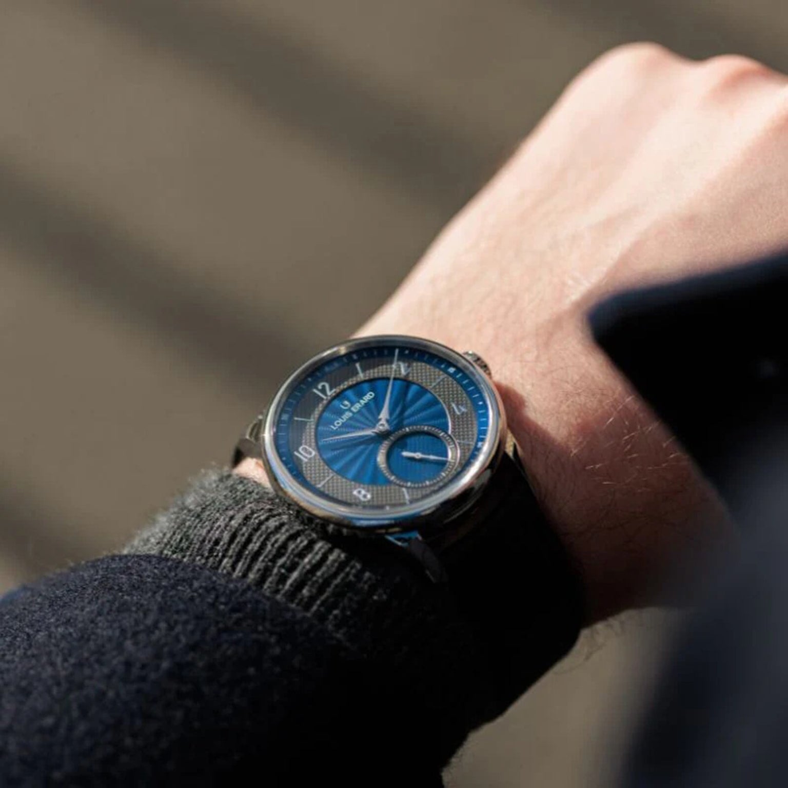 A person wearing the Louis Erard Petite Seconde Guilloché Anthracite Blue wristwatch, featuring a blue face with silver details, is seen outdoors in natural light.