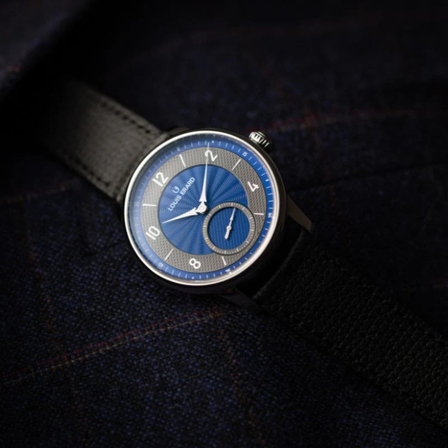 The Louis Erard Petite Seconde Guilloché Anthracite Blue wristwatch, featuring a blue dial and black strap, is displayed on a dark fabric background.