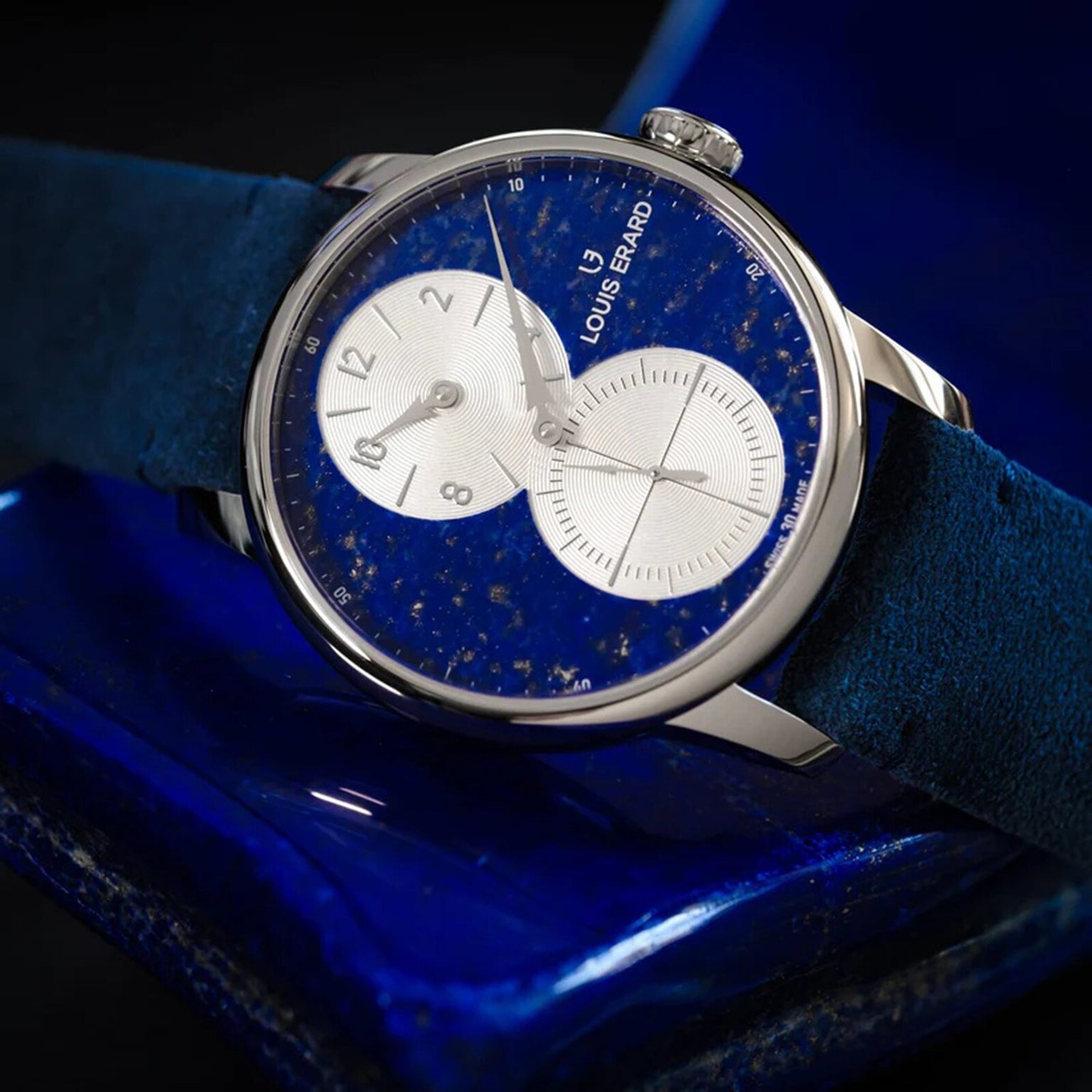 The Louis Erard Régulateur Lapis-Lazuli wristwatch features a striking blue face with dual subdials, shown here resting on a dark blue surface.