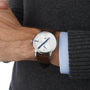 A person wearing the Louis Erard Le Régulateur Louis Erard x seconde/seconde/, a silver wristwatch with a white face and brown leather strap, over a gray sweater.