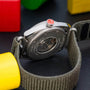 Close-up of the Louis Erard Le Régulateur x Alain Silberstein Khaki wristwatch back with a grey fabric strap, surrounded by colorful wooden blocks.
