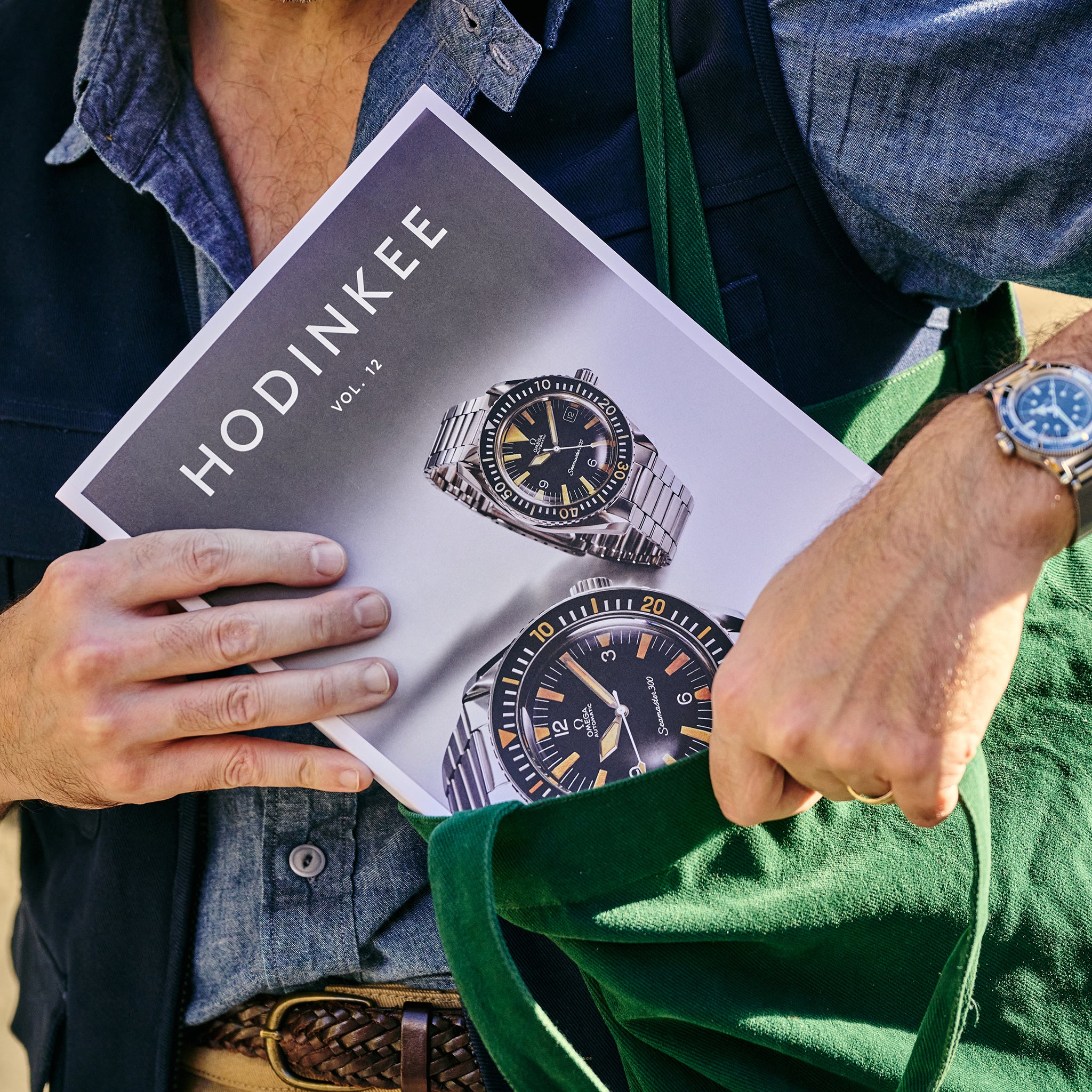 A person, wearing a watch on their left wrist, slides the HODINKEE Magazine, Volume 12 by Hodinkee—featuring watches on the cover—partially into a green bag, capturing vintage watch style.