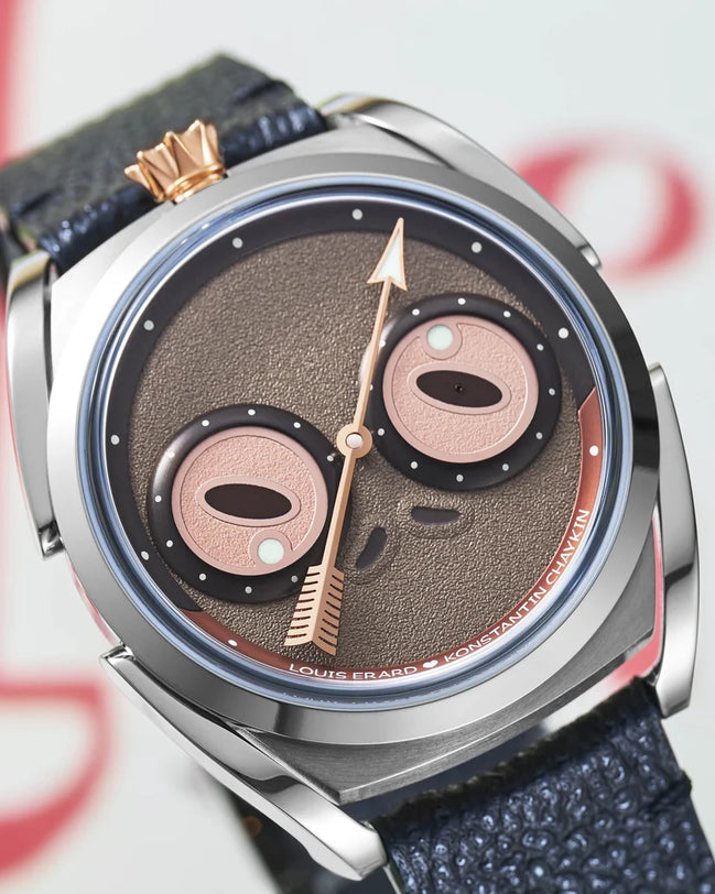 Titanium watch with brown textured dial featuring two pink circular eyes, gold hands, and dark blue leather strap with gold c