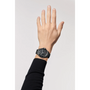 A raised left hand wears a Chanel J12 GMT wristwatch with a round face, high-precision white hour markers, and a black band. The wearer has on a long-sleeve black shirt against a plain, light-colored background.