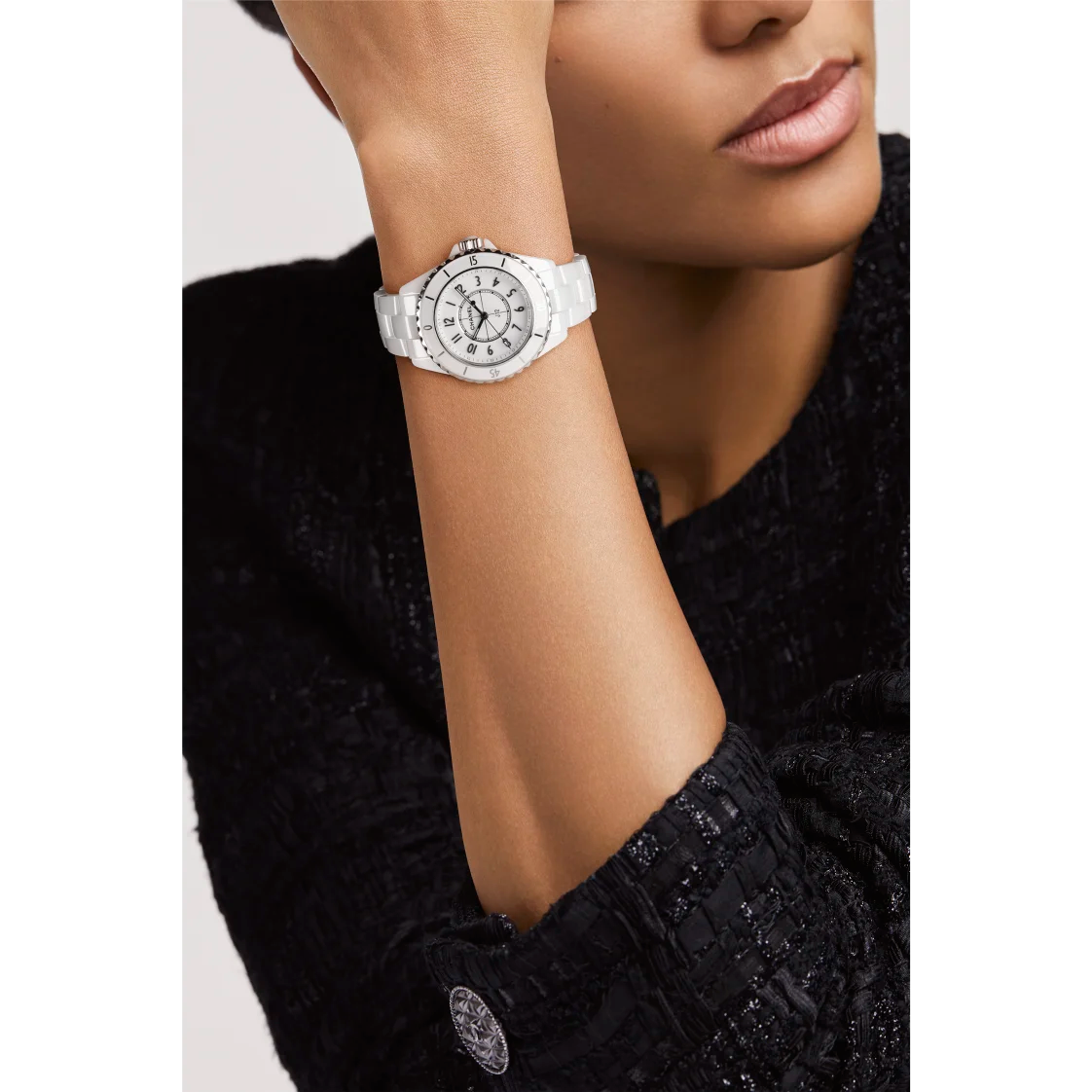A person in a textured black top rests their chin on their hand, showing a Chanel J12 wristwatch with a white round face and elegant silver accents.