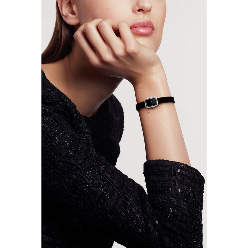 A woman in a textured black long-sleeve top rests her chin on her hand, showcasing the Chanel Première Ribbon—a small, square wristwatch with a black-lacquered dial and black strap—against a plain white background.