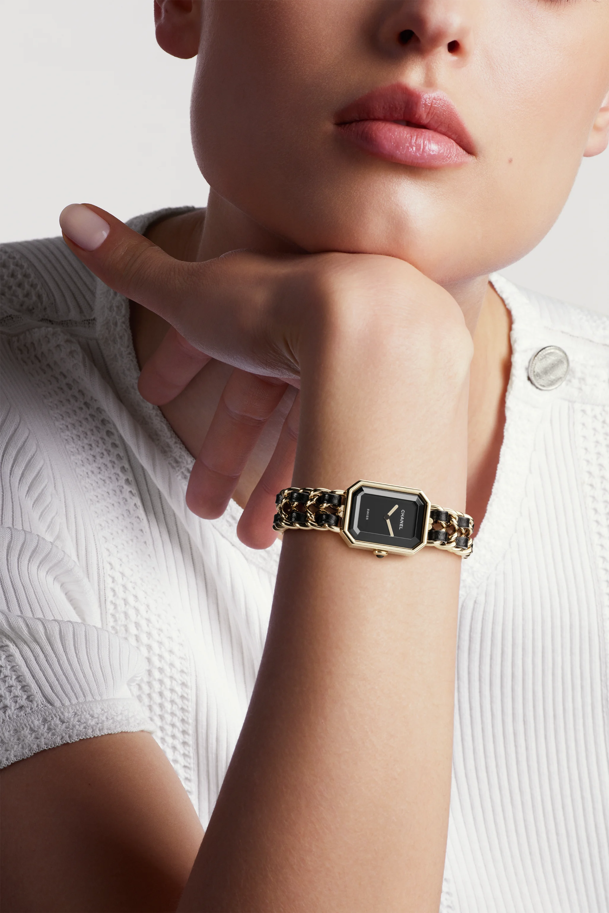 A person in a textured white top rests their chin on their hand, showcasing the Chanel Première Édition Originale Small Model wristwatch with a black-lacquered square dial and chain-link band.