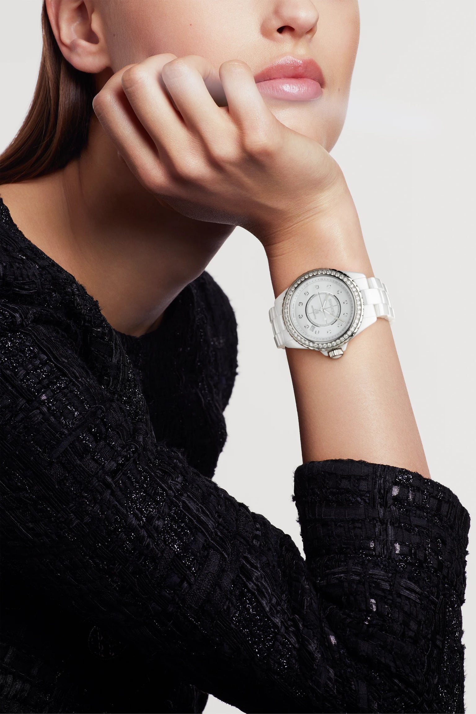 A person in a textured black top rests their chin on their hand, showcasing the Chanel J12 Diamond Bezel—a white wristwatch with a durable ceramic and steel case and a round, crystal-studded bezel—against a plain, light background.