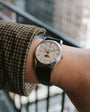 A person stands by a railing, wearing the Patek Philippe Retrograde Perpetual Calendar—a silver wristwatch with a white dial and black leather strap.