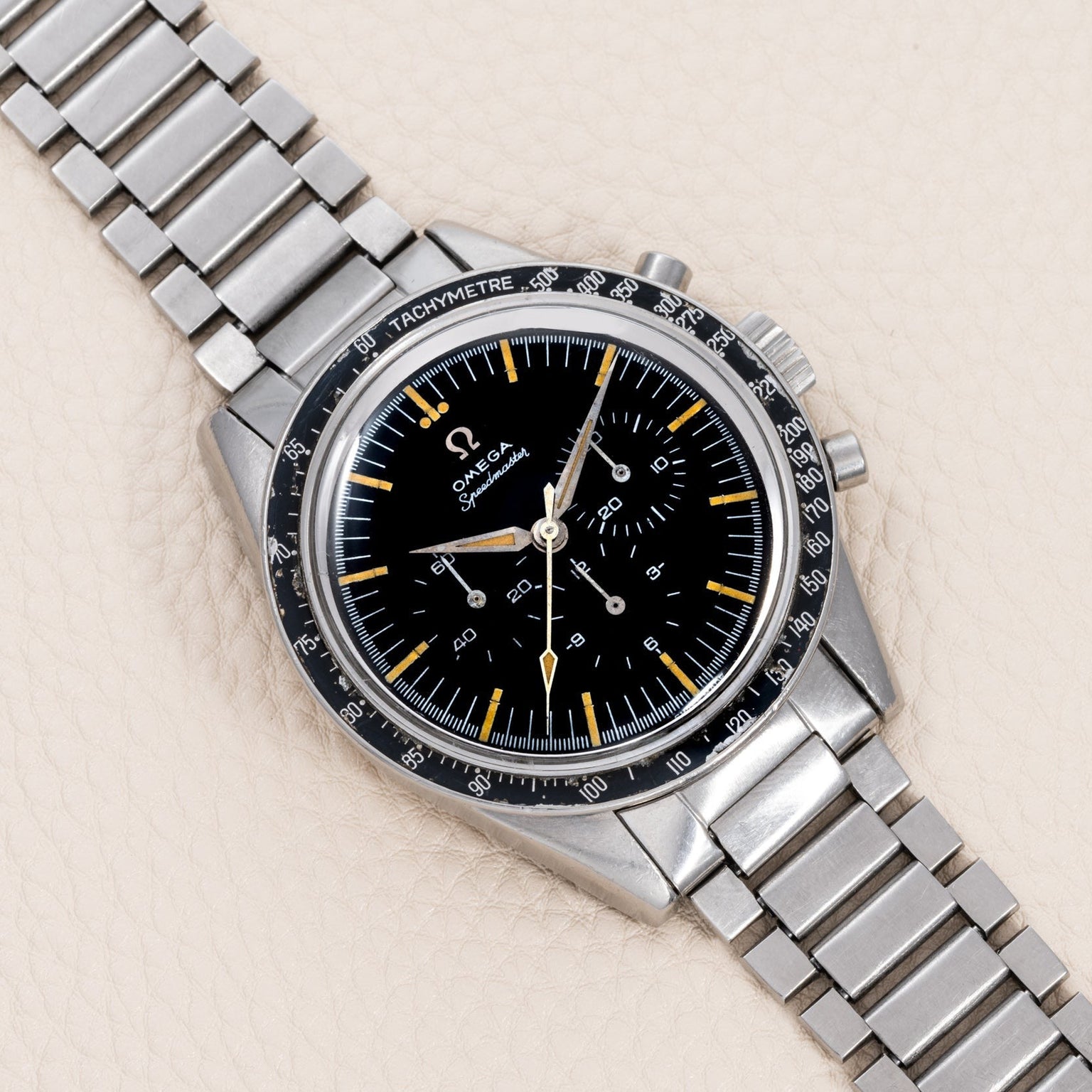 An OMEGA Speedmaster watch with a black dial and metal bracelet displayed on a beige textured surface.