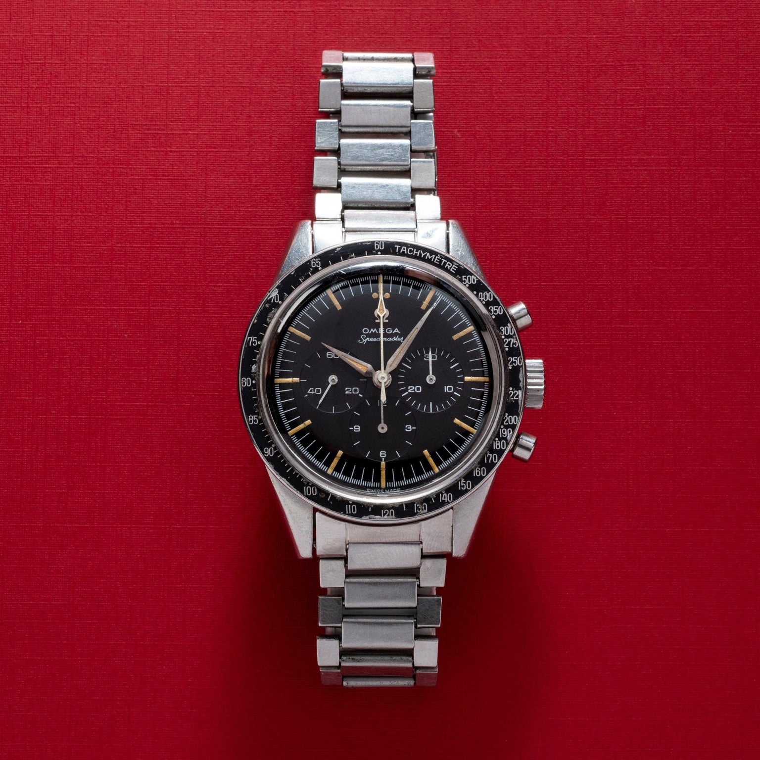 A silver OMEGA Speedmaster wristwatch with a black dial displayed on a red textured background.