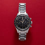 A silver OMEGA Speedmaster wristwatch with a black dial displayed on a red textured background.