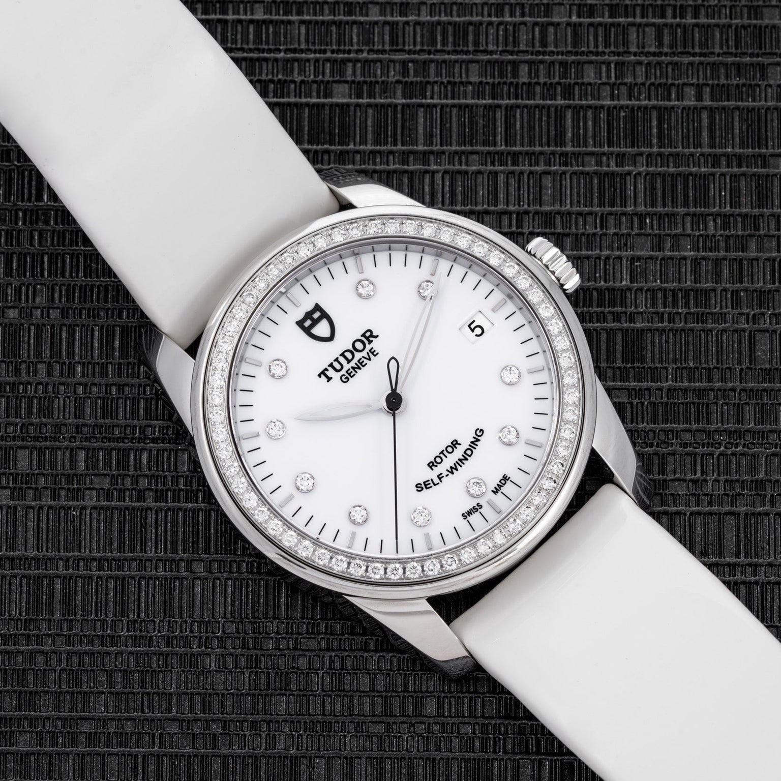 The TUDOR Glamour Date wristwatch features a white strap and diamond bezel, showcased on a textured black surface.