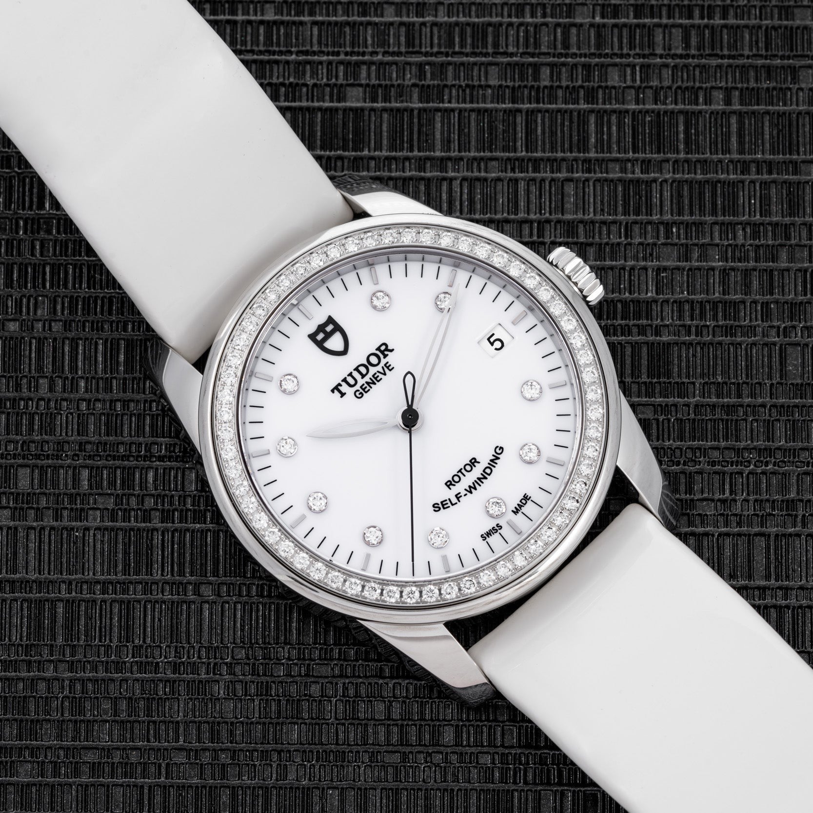 The TUDOR Glamour Date wristwatch features a white strap and diamond bezel, showcased on a textured black surface.