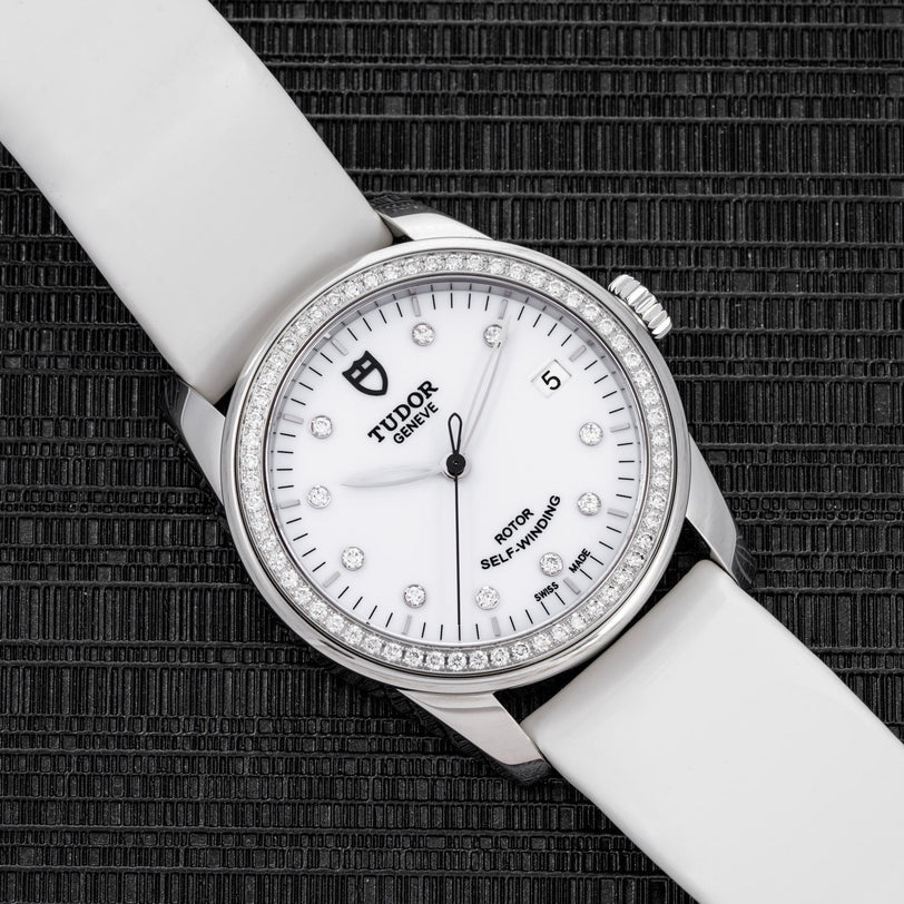 The TUDOR Glamour Date wristwatch features a white strap and diamond bezel, showcased on a textured black surface.