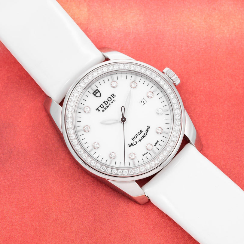 The TUDOR Glamour Date watch with a white strap and diamond markers is displayed on a red textured background.