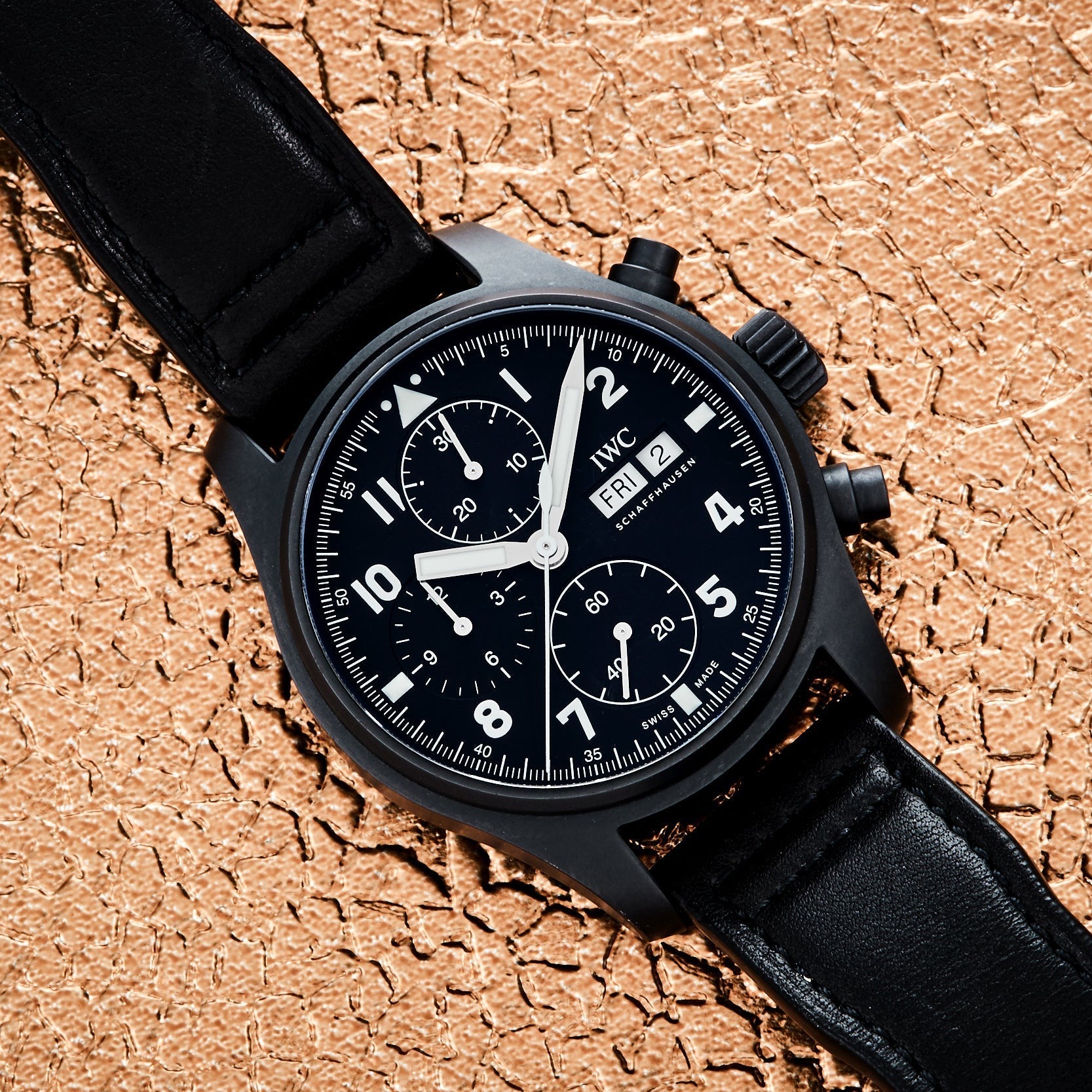 The IWC Schaffhausen Pilots Watch Chronograph Tribute To 3705 with a black dial and leather strap is displayed on a textured beige surface.