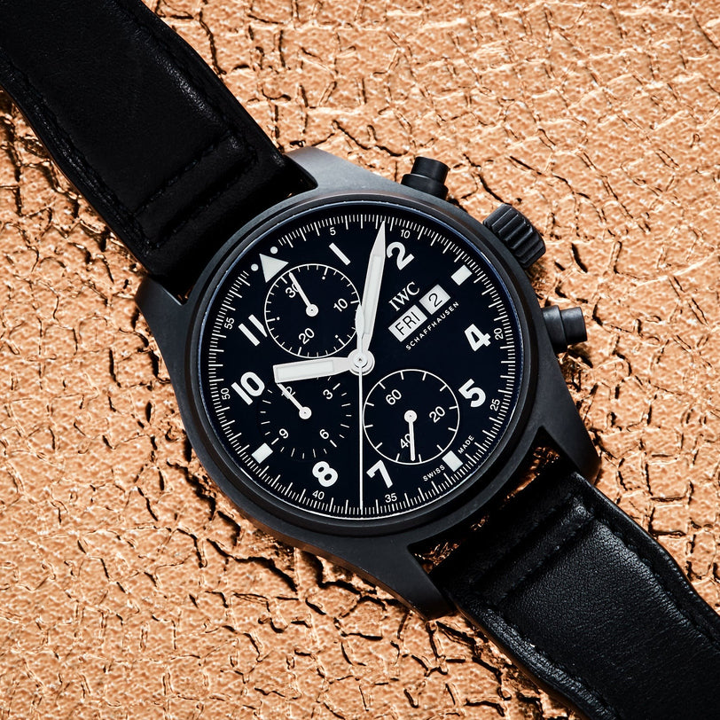 The IWC Schaffhausen Pilots Watch Chronograph Tribute To 3705 with a black dial and leather strap is displayed on a textured beige surface.
