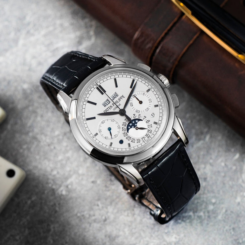 The Patek Philippe Perpetual Calendar Moonphase Chronograph is a luxury wristwatch featuring a black leather strap, white dial, multiple subdials, and date indicators.