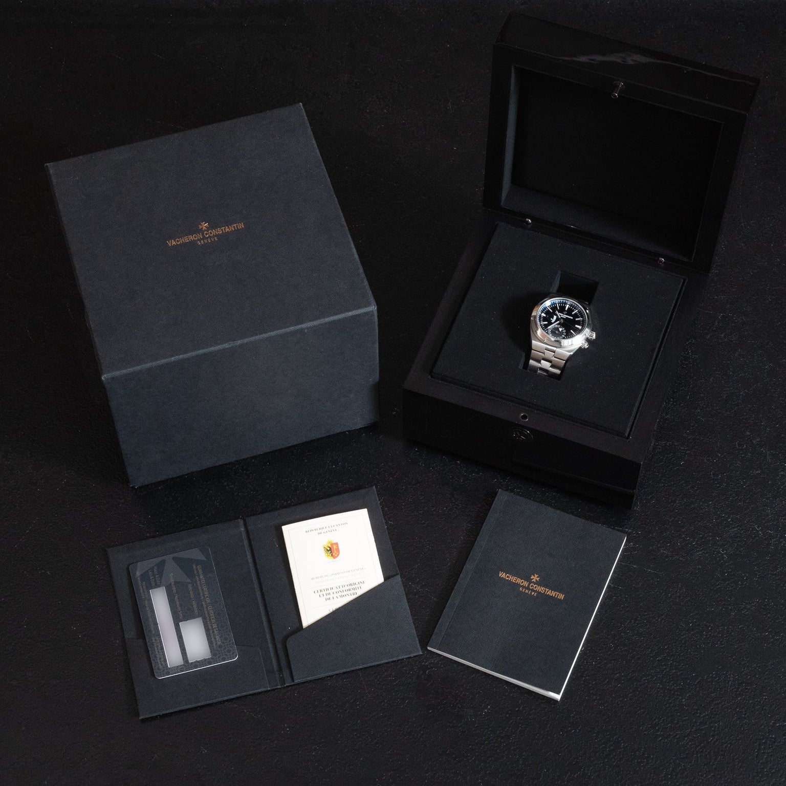 The Vacheron Constantin Overseas Dual-Time luxury watch is presented in its original black box with warranty cards, booklet, and packaging, all elegantly displayed on a dark surface.