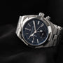 The Vacheron Constantin Overseas Dual-Time, a luxury silver wristwatch with a blue dial and metal band, is showcased on a black fabric background.