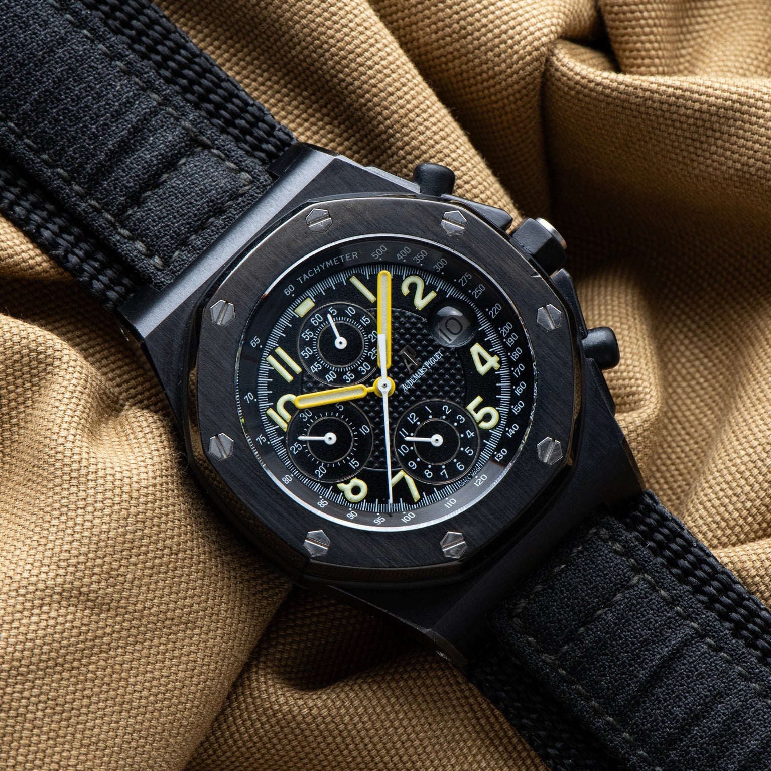 The Audemars Piguet Royal Oak Offshore End of Days chronograph watch features yellow accents on a black strap, displayed on tan fabric.