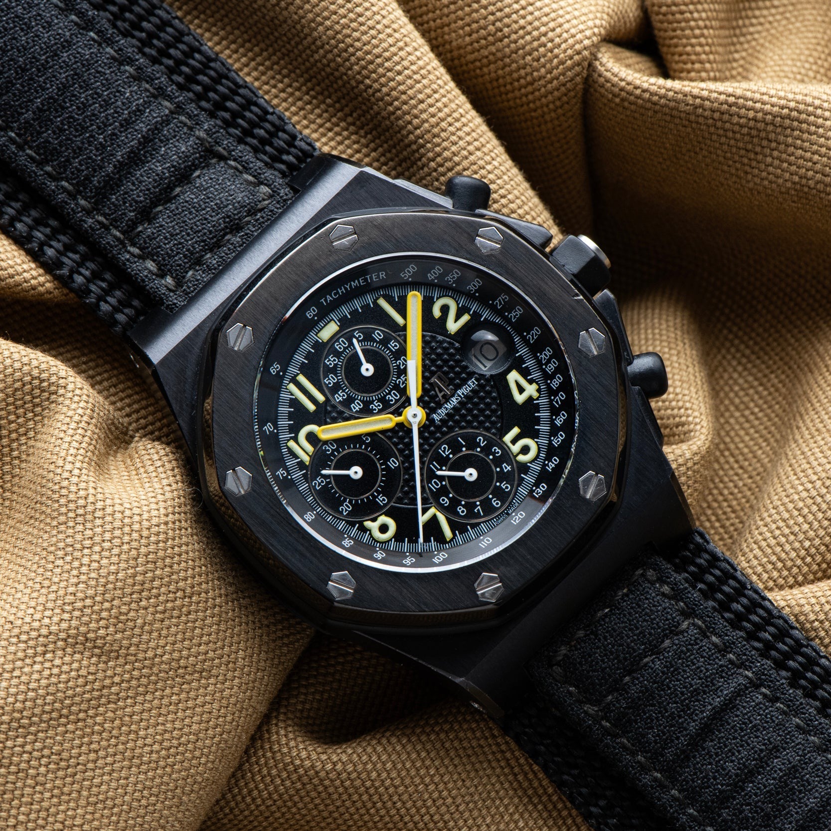 The Audemars Piguet Royal Oak Offshore End of Days chronograph watch features yellow accents on a black strap, displayed on tan fabric.